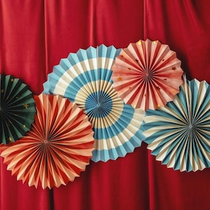 May include: A collection of colorful paper fans in various sizes and designs. The fans feature shades of red, blue, green, and cream, with star accents on some. They are arranged against a red curtain backdrop, creating a festive and decorative display.