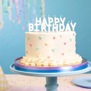 Op de afbeelding: Een crème verjaardagstaart met witte 'HAPPY BIRTHAY'-kaarsen. De taart is versierd met pastelkleurige hartjes en staat op een taartstandaard met blauwe en paarse linten. Feestmutsen staan op de achtergrond.