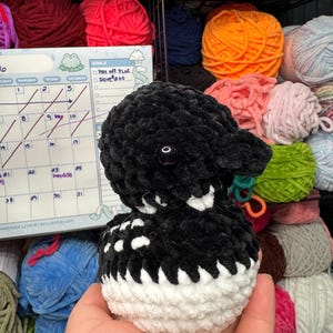 May include: A handcrafted black and white plush toy, possibly a bird, with a soft, fluffy texture. The toy features a white belly and a black head and back. The background includes colourful yarn skeins and a calendar.