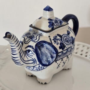 May include: A ceramic teapot shaped like an elephant, painted in white and blue. The teapot features intricate floral and geometric patterns, with a curved spout and a handle. The lid is topped with a small finial. The teapot is on a white plate.