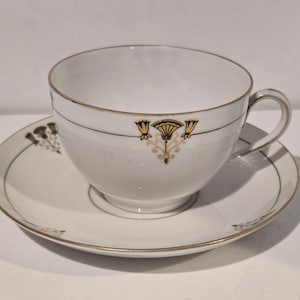 May include: White teacup and saucer set with gold trim and floral accents. The teacup has a handle and is decorated with a gold and black floral design. The saucer has a matching design. A classic tea set.