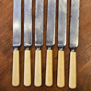 May include: Set of six vintage butter knives. Each knife features a stainless steel blade and a cream-colored handle. The knives are arranged on a dark wooden surface, showcasing their classic design and potential for use in a formal dining setting.