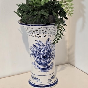 Könnte beinhalten: Eine weiß-blaue Keramikvase mit floralem Design, gefüllt mit grünen Kunstpflanzen. Die Vase hat einen ausgestellten oberen Rand mit dekorativen Aussparungen und einen blauen Rand. Das florale Design ist in Blautönen gehalten.