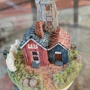 Puede incluir: Un faro en miniatura de cerámica con dos pequeñas casas, una roja y otra azul, con vallas blancas y una base de hierba verde.