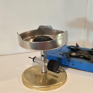 May include: A vintage camping stove with a silver burner and a brass base. The stove has a black control knob and a small, black burner. A blue stove is in the background.