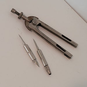 Puede incluir: Un compás vintage de plata con dos puntas intercambiables, una con una punta afilada y la otra con una pluma.