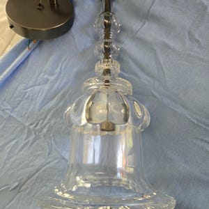 May include: Clear glass pendant light with a decorative, bell-shaped shade. The fixture has a metal canopy and a black cord. The shade has a scalloped edge and a decorative stem. The light is designed for indoor use.