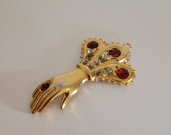 Könnte beinhalten: Ein goldfarbener Anhänger mit einer ausgestreckten Hand, die eine rote Edelstein auf der Fingerspitze trägt. Die Hand ist an ein kronenartiges Design mit drei roten Edelsteinen und klaren Strasssteinen befestigt.
