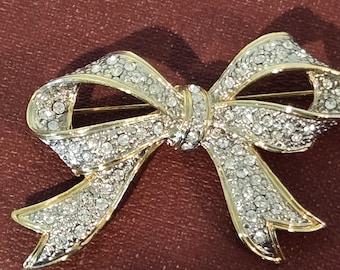 May include: A gold-toned bow brooch encrusted with clear crystals. The bow design features a detailed knot in the center and gracefully curved loops. The brooch is set against a dark red-brown background.