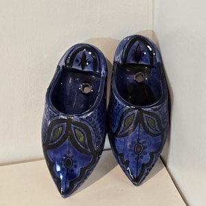 May include: Two blue ceramic clogs with decorative floral patterns in black and green. The clogs have a glossy finish and a hole at the top. The clogs are displayed on a white surface.