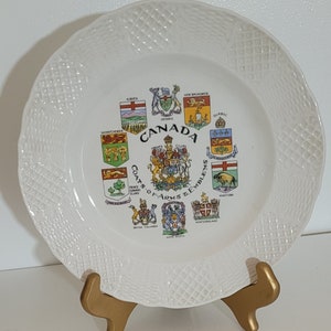 May include: White decorative plate featuring the coats of arms and emblems of Canadian provinces. The plate has a textured rim and is displayed on a gold-colored stand. The word "CANADA" is prominently displayed.