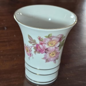 以下が含まれることがあります： 上部がフレアになった白いセラミック花瓶、金色の縁取り。ピンクと黄色の花と緑の葉のフローラルデザイン。金色のバンドが花瓶の底を囲んでいます。