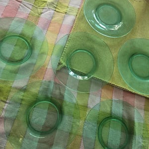 Puede incluir: Colección de seis platos de vidrio transparente de color verde. Los platos son redondos con un centro circular en relieve. Los platos están colocados sobre un mantel a rayas de colores y un paño verde.