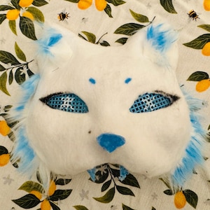 Op de afbeelding: Een wit kattenmasker met blauwe details. Het masker heeft blauwe mesh oogbedekkingen, een blauwe neus en blauwe bontafwerking. Gemaakt van een zacht, viltachtig materiaal. De achtergrond is een bloemenpatroon.
