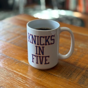 May include: A white ceramic mug with a handle, featuring the text "KNICKS IN FIVE" printed in blue block letters with an orange outline. The mug is filled with a dark liquid, likely coffee, and has a smooth, glossy finish on its exterior surface.