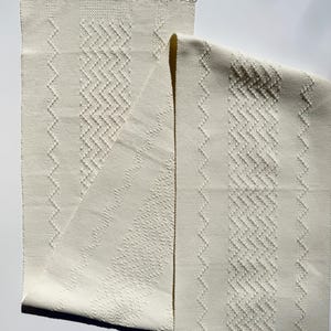 May include: Off-white woven textile with geometric patterns and fringe. The textile features zig-zag and vertical line designs. The fringe is at the top. The textile is folded over on itself. The measurements are in centimetres.