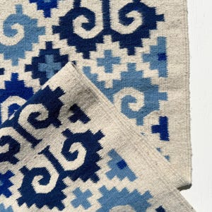 May include: A woven rug with a geometric pattern in shades of blue and white. The rug has a fringed edge and is made of natural fibres.