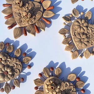 May include: Close-up of several beige clay heart-shaped wall decorations. Each heart features floral and leaf designs, with small brown accents and a wire backing. The hearts are arranged on a white background, casting shadows.