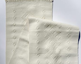 Op de afbeelding: Crème geweven textiel met een gefranjerde rand. Het textiel heeft een geometrisch patroon van ruiten en verticale strepen. De stof is gevouwen en onthult een donkerblauwe rand aan de onderkant. De franje is crème.