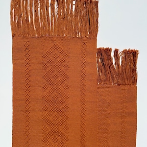 May include: Two rust-coloured woven textile pieces with fringed edges. The larger piece features a diamond pattern and vertical stripes. The smaller piece has a similar design. The textiles are likely handmade and suitable for home decor.