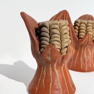 May include: Two terracotta-colored ceramic corn sculptures. Each sculpture features a detailed ear of corn nestled within a stylized, petal-like base. The base has vertical, light-colored stripes.