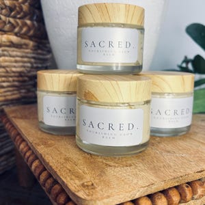 Puede incluir: Cuatro frascos de vidrio con tapas de madera que contienen un bálsamo de color crema. Los frascos están apilados en una bandeja de madera con un borde de cuentas. Los frascos están etiquetados con "SACRED. NOURISHING GLOW BALM."