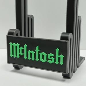Op de afbeelding: Een zwart en grijze standaard met het woord "McIntosh" in groene letters. De standaard heeft een rechthoekige basis en een reeks gebogen, verticale steunen. De belettering is in een gotisch lettertype.