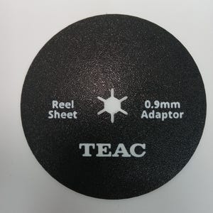 May include: A black, circular TEAC reel sheet adaptor with white text. The text includes "Reel Sheet", "0.9mm Adaptor", and the brand name "TEAC". A white, six-pointed star is in the center.