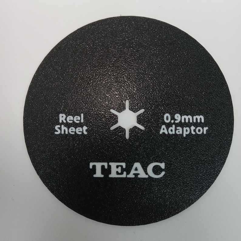 May include: A black, circular TEAC reel sheet adaptor with white text. The text includes "Reel Sheet", "0.9mm Adaptor", and the brand name "TEAC". A white, six-pointed star is in the center.