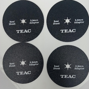 May include: Four black circular reel sheet adaptors. Each adaptor has the text "Reel Sheet", a star-shaped design, "0.9mm Adaptor", and the brand name "TEAC" in white text. The adaptors are designed for audio equipment.