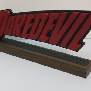 Op de afbeelding: Een rood en zwart 3D-geprint bord met het woord "DAREDEVIL" in vetgedrukte letters. Het bord is gemonteerd op een houten basis.