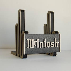 May include: A black and gold display stand featuring a textured rectangular front panel with the word McIntosh embossed in a gothic-style font. The structure has two vertical, layered, wave-like side supports with gold-toned edges.