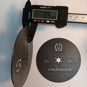 May include: A digital caliper measures a black disc with a 0.9 mm thickness. The disc has the word "PIONEER" printed on it, along with the text "Reel Sheet" and "0.9mm Adaptor".