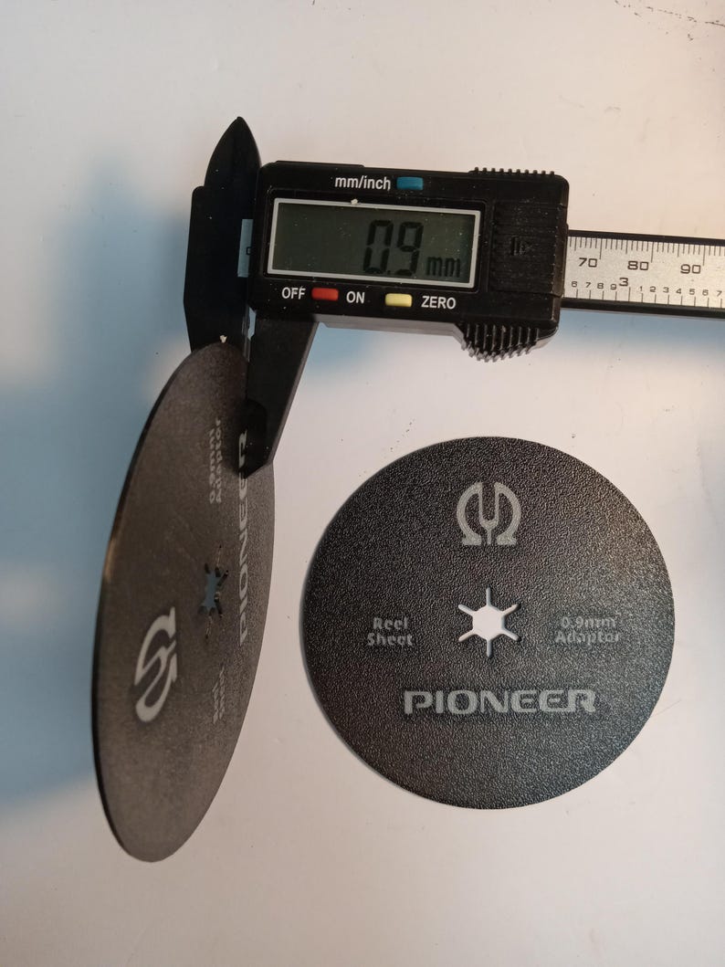 May include: A digital caliper measures a black disc with a 0.9 mm thickness. The disc has the word "PIONEER" printed on it, along with the text "Reel Sheet" and "0.9mm Adaptor".