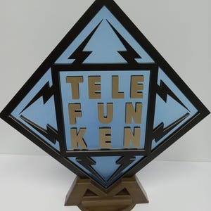 Puede incluir: Una placa negra en forma de diamante con fondo azul y letras doradas que dicen "TELEFUNKEN". La placa está montada en un soporte de madera marrón.