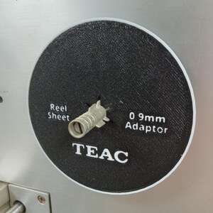 May include: Close-up of a black circular component with white text that reads "Reel Sheet" and "0.9mm Adaptor". The word "TEAC" is also printed in white. A silver metal adaptor is in the center of the component. The background is a silver metal surface.