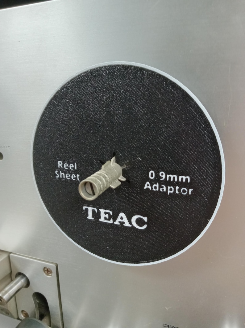 May include: Close-up of a black circular component with white text that reads "Reel Sheet" and "0.9mm Adaptor". The word "TEAC" is also printed in white. A silver metal adaptor is in the center of the component. The background is a silver metal surface.