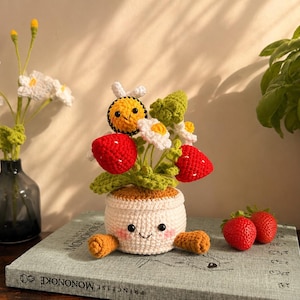 Peut inclure: Une plante en crochet dans un pot blanc avec un visage souriant, des joues roses et deux bras marron. La plante comprend des fraises rouges, des marguerites blanches, des feuilles vertes et une petite abeille jaune et noire avec des ailes blanches.