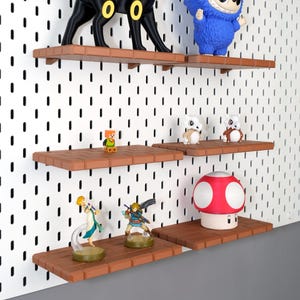 May include: A display of figurines on brown shelves against a white pegboard. The figurines include a black Umbreon, a blue character, a Minecraft figure, two white and brown figures, a Zelda figurine, a Link figurine, and a red and white mushroom.