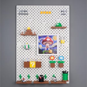 Puede incluir: Un tablero perforado blanco con varios accesorios temáticos de Super Mario. Incluye una impresión enmarcada de un hongo al estilo de La noche estrellada de Van Gogh, junto con estantes, figuras y el texto "MARIO" y "TIME 999".