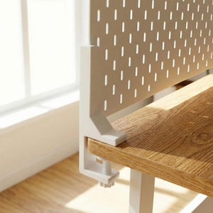 Puede incluir: Un organizador de escritorio de metal blanco con un panel trasero perforado y una abrazadera para sujetar a un escritorio. El escritorio tiene un acabado de veta de madera clara. El organizador está diseñado para guardar material de oficina.