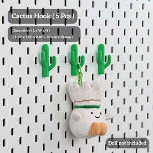 Puede incluir: Ganchos verdes con forma de cactus sobre una pared perforada blanca. Los ganchos miden aproximadamente 3,5 cm de largo, 3 cm de ancho y 6,8 cm de alto. El texto "Cactus Hook (5 Pcs)" es visible. Una mu&ntilde;eca de peluche cuelga de uno de los ganchos.