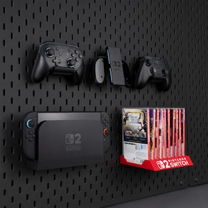 May include: A black pegboard wall mount system holding a Nintendo Switch console, two wireless controllers, a charging grip, and a row of game cases on a red shelf with the white text NINTENDO SWITCH printed on the front edge of the shelf.