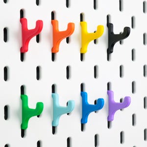May include: A set of eight Y-shaped pegboard hooks in red, orange, yellow, black, green, light blue, blue, and purple, each featuring a dual-prong design for hanging items, mounted onto a white pegboard with vertical slots for organization.