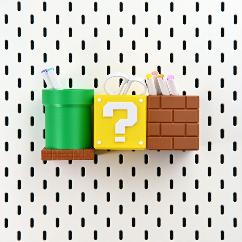 May include: A set of three decorative storage containers inspired by Super Mario Bros. The containers are a green pipe, a yellow question mark block, and a brown brick block. They hold pens, scissors, and markers, ideal for a desk.