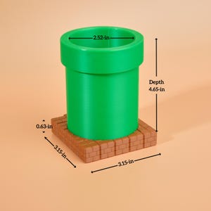 May include: A green pipe with a 6.4 cm diameter and a 11.8 cm depth, set on an 8 cm square brick base. The base is 1.6 cm thick. The pipe is a vibrant green colour, and the base is a brown brick colour.