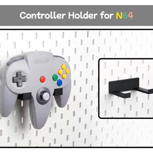 May include: A black plastic controller holder for N64 mounted on a white pegboard. The holder features two horizontal prongs to support a grey Nintendo 64 game controller. The text Controller Holder for N64 is displayed at the top of the image.