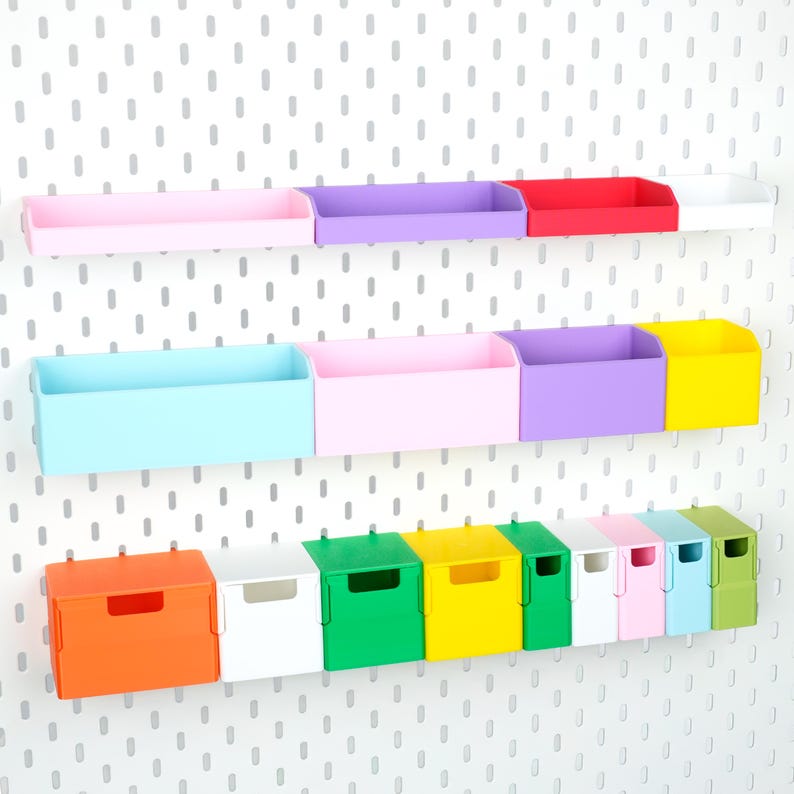 May include: A collection of colourful plastic storage bins and containers mounted on a white pegboard. The set includes various rectangular trays and small square boxes in shades of pink, purple, red, white, blue, yellow, orange, and green.