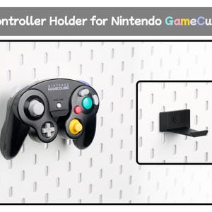 May include: A black plastic controller holder for a Nintendo GameCube console, designed to mount onto a white pegboard. The holder features a small embossed logo and is shown both empty and supporting a black Nintendo GameCube game controller.