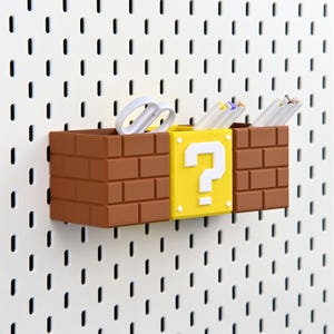 May include: A desk organiser in the style of a Super Mario Bros. level, featuring brown brick-like containers and a yellow question mark block. White scissors and pencils are stored inside. The organiser is mounted on a white pegboard.